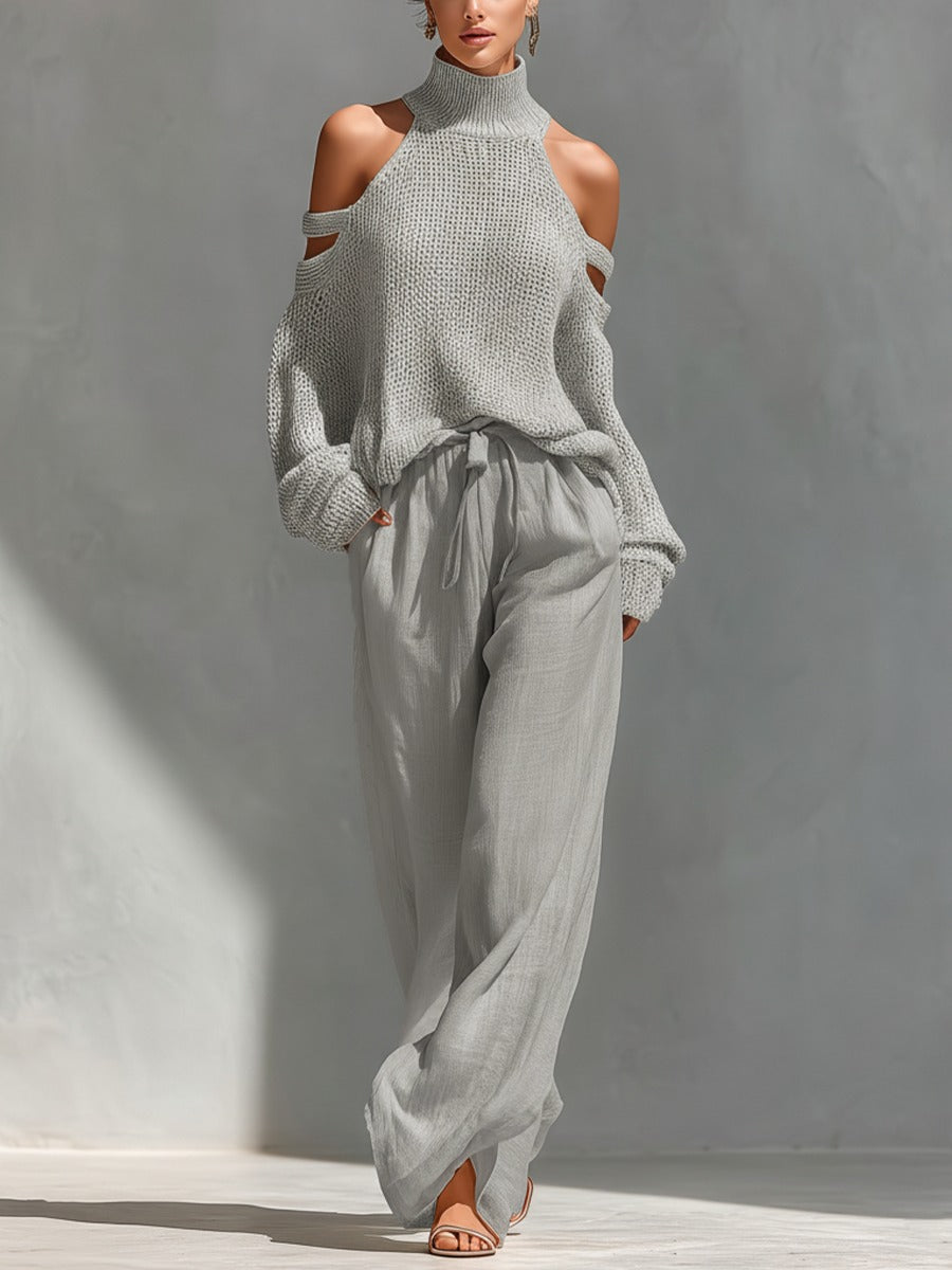 Wearing the rosalie | Ensemble Deux Pièces Off-Shoulder in light gray, a person stands against a softly lit gray background, showcasing an elegant two-piece look with a knit sweater and loose, high-waisted pants.