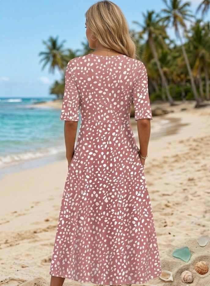 A woman with long blonde hair stands on a sandy beach, facing away, wearing the Luna | ROBE MIDI À POIS ÉLÉGANTE ET FLATTEUSE POUR LE VENTRE. Palm trees and the ocean are behind her, with seashells scattered on the sand.