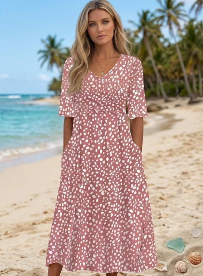 A woman with long blonde hair wears the Luna | ROBE MIDI À POIS ÉLÉGANTE ET FLATTEUSE POUR LE VENTRE in pink and white polka dots, standing on a sandy beach with palm trees, ocean views, and seashells around her feet.